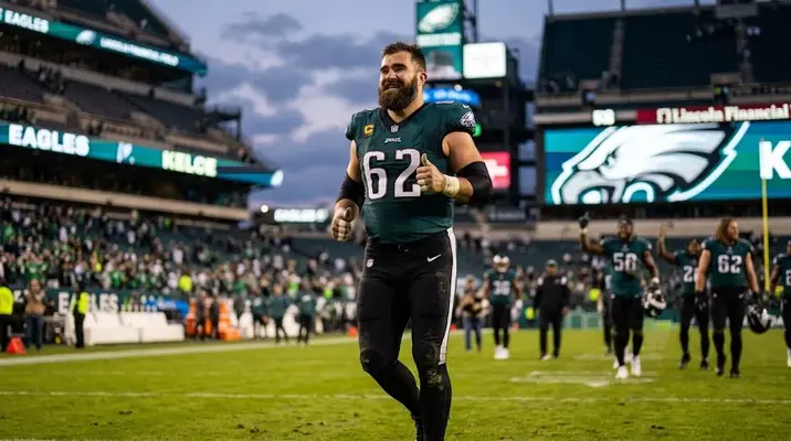 Jason Kelce smiling in a suit at 'The Philadelphia Center' Eagles event, '62' eagle lapel pin, hands clasped, warm indoor lighting.